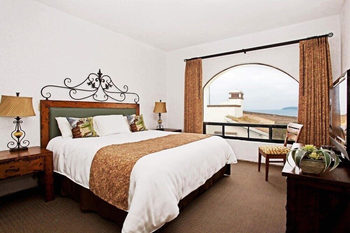 A hotel room with a bed, two bedside tables with lamps, a desk with a TV, and a large window with curtains.