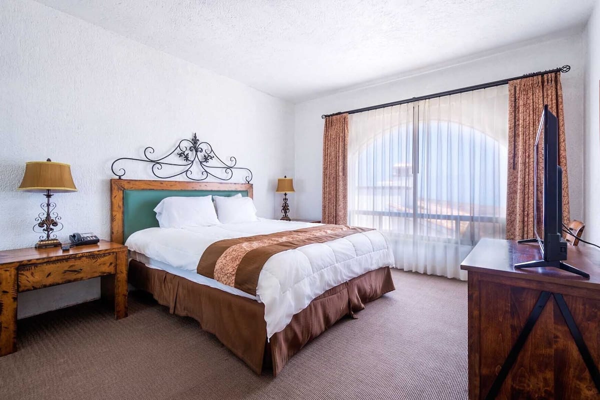 A hotel room with a bed, two bedside tables, a desk, and a large window with curtains.
