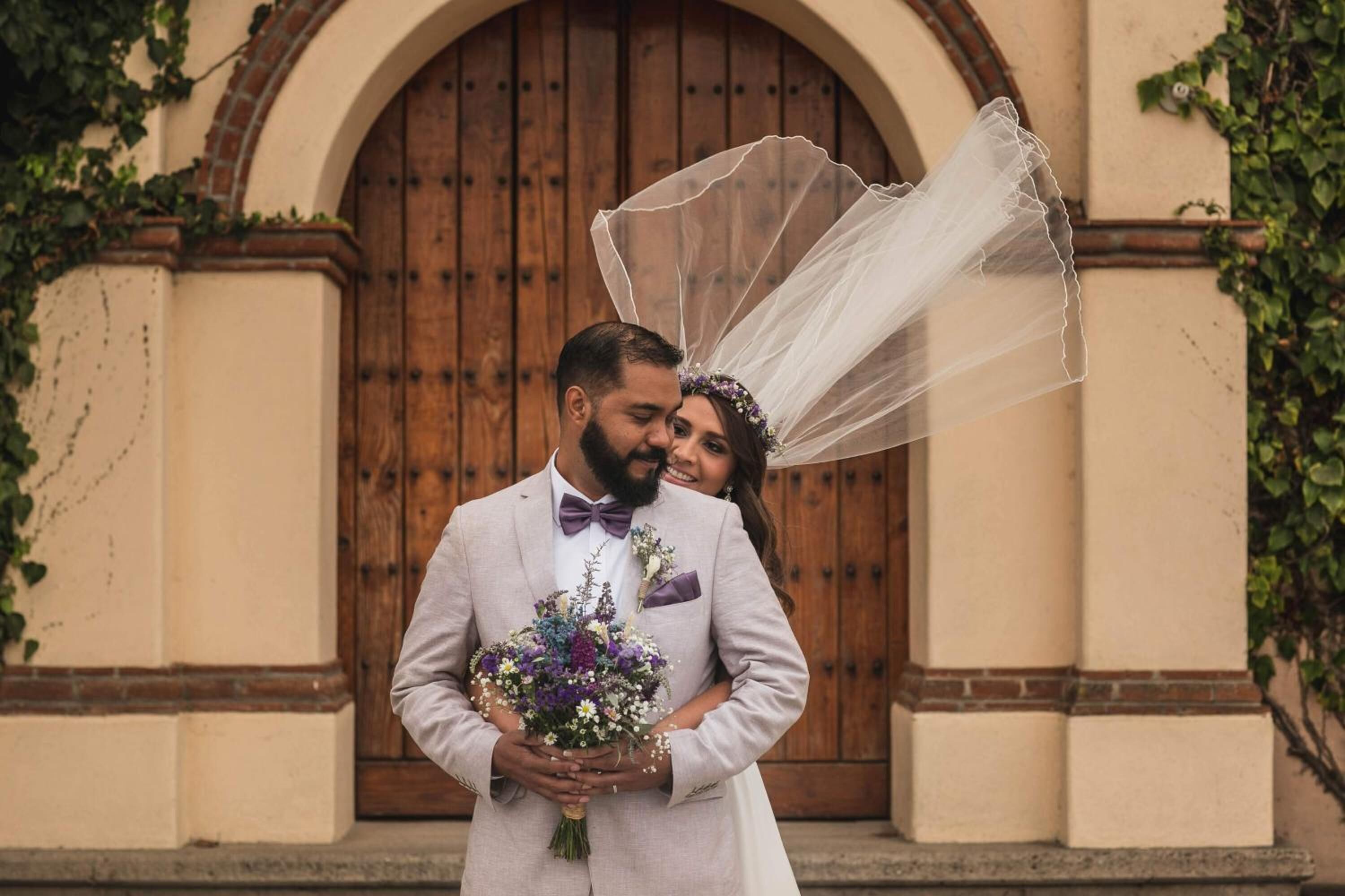 Una pareja abrazándose frente a una puerta de madera grande, con una pared color beige y follaje a los lados.