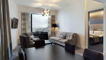 A modern hotel room with a large window, a sofa, a dining table, and a separate sleeping area.