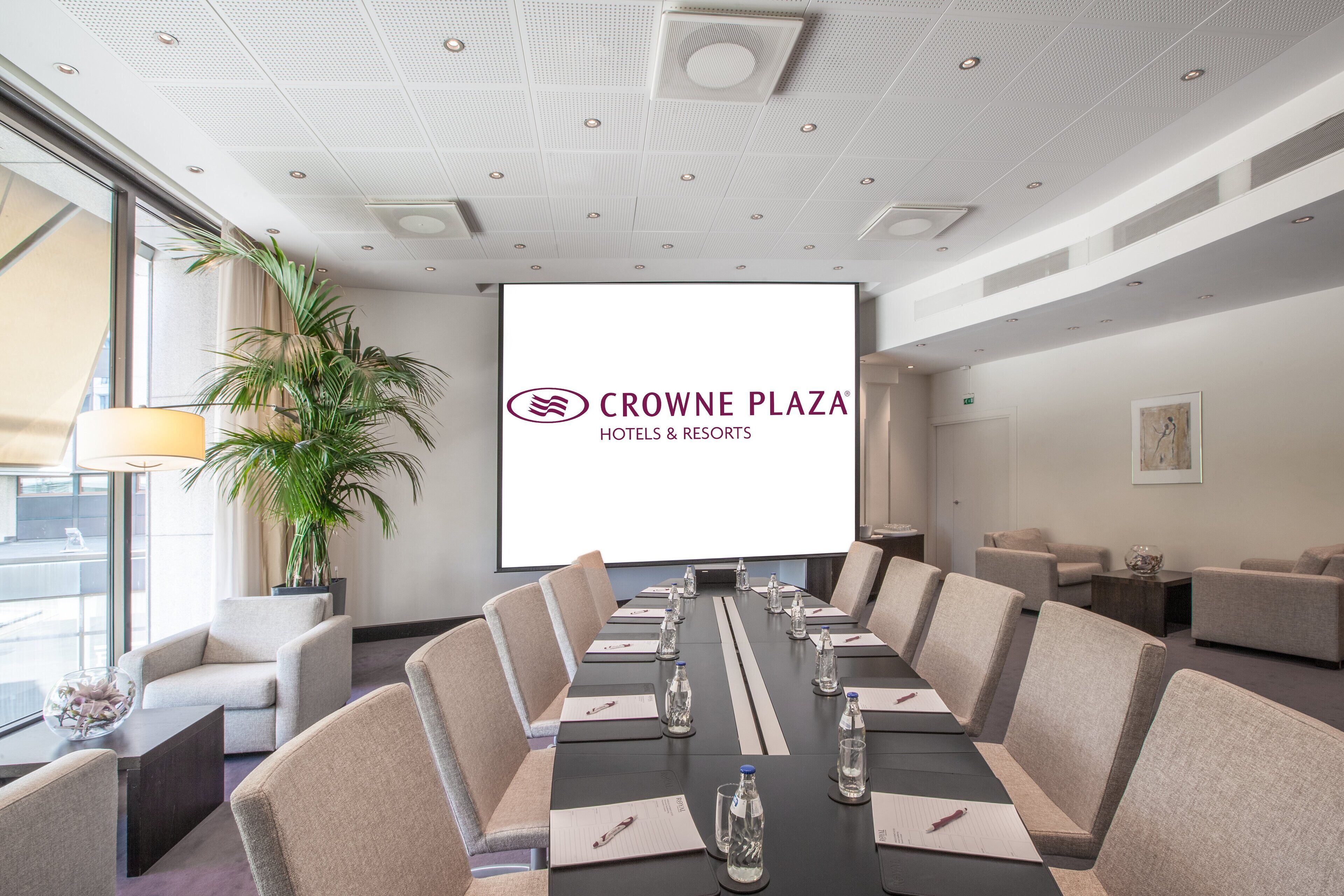 Kokoushuone, jossa on pitkä pöytä, beigenväriset tuolit, projektorikangas, jossa näkyy teksti ”CROWNE PLAZA HOTELS & RESORTS”, ja ruukussa oleva viherkasvi.