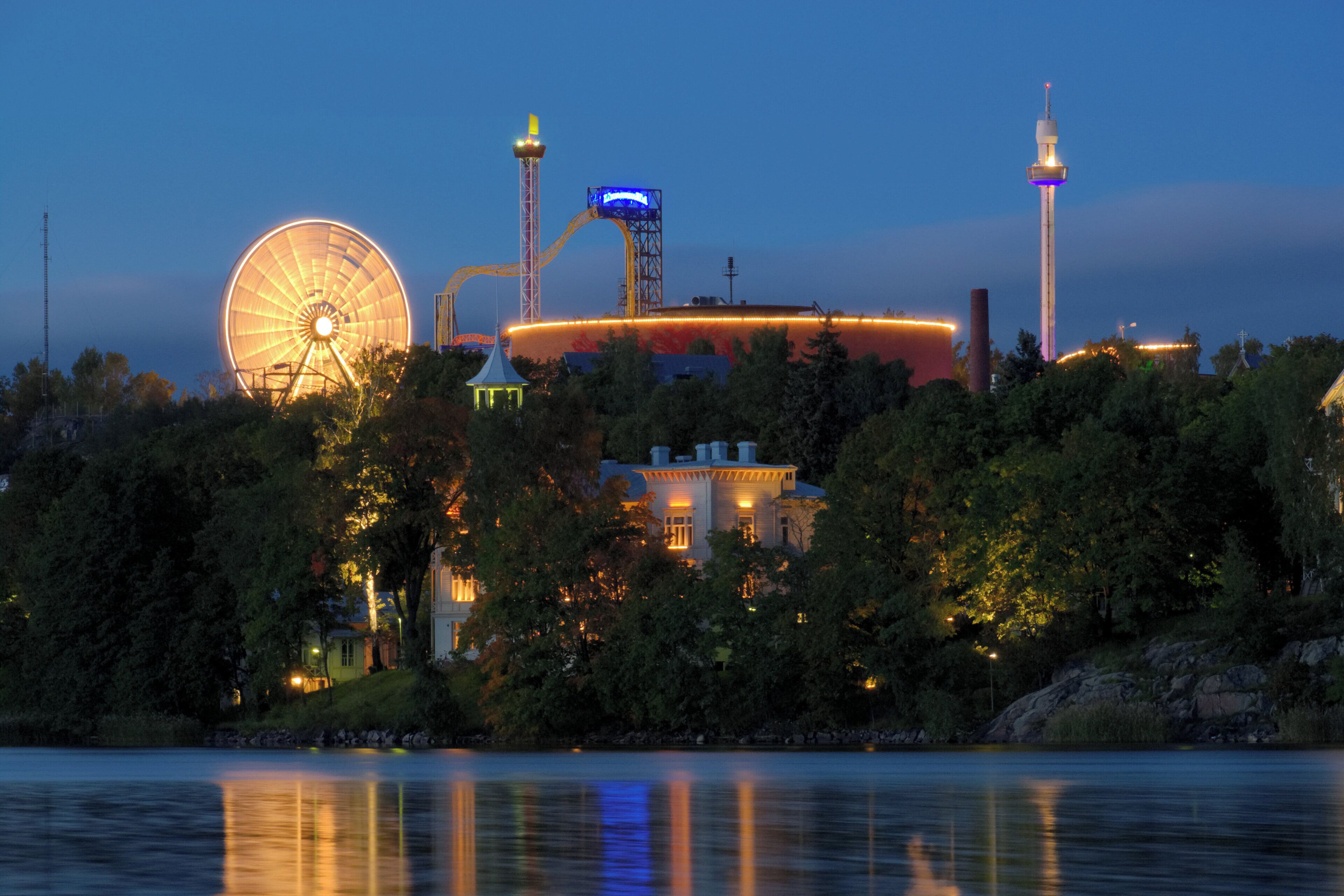A waterfront view with a large Ferris wheel, a building with a tower, and a body of water reflecting the structures.