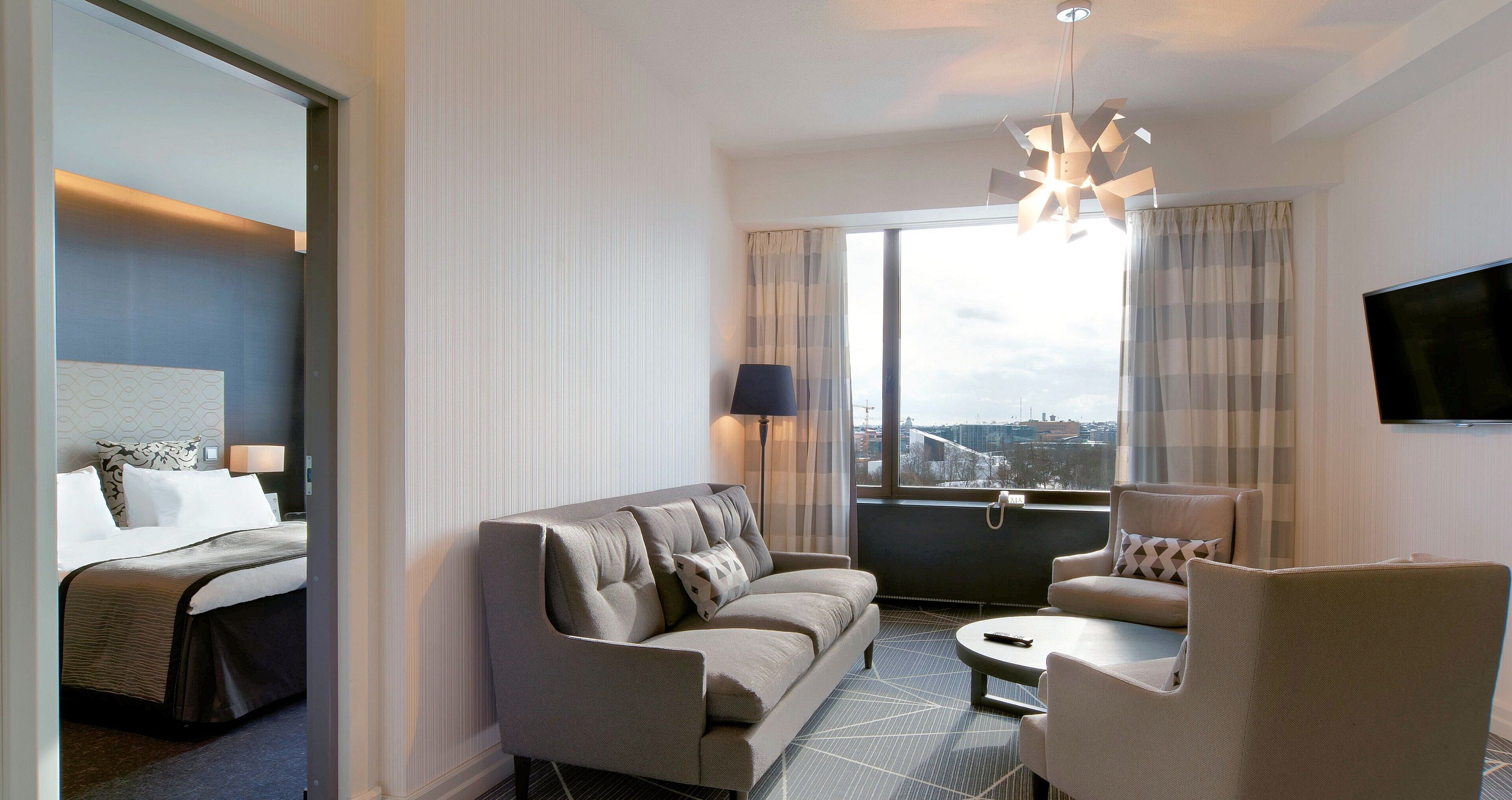A hotel room with a bed, a sofa, a chair, a small table, a TV, and a large window with curtains.