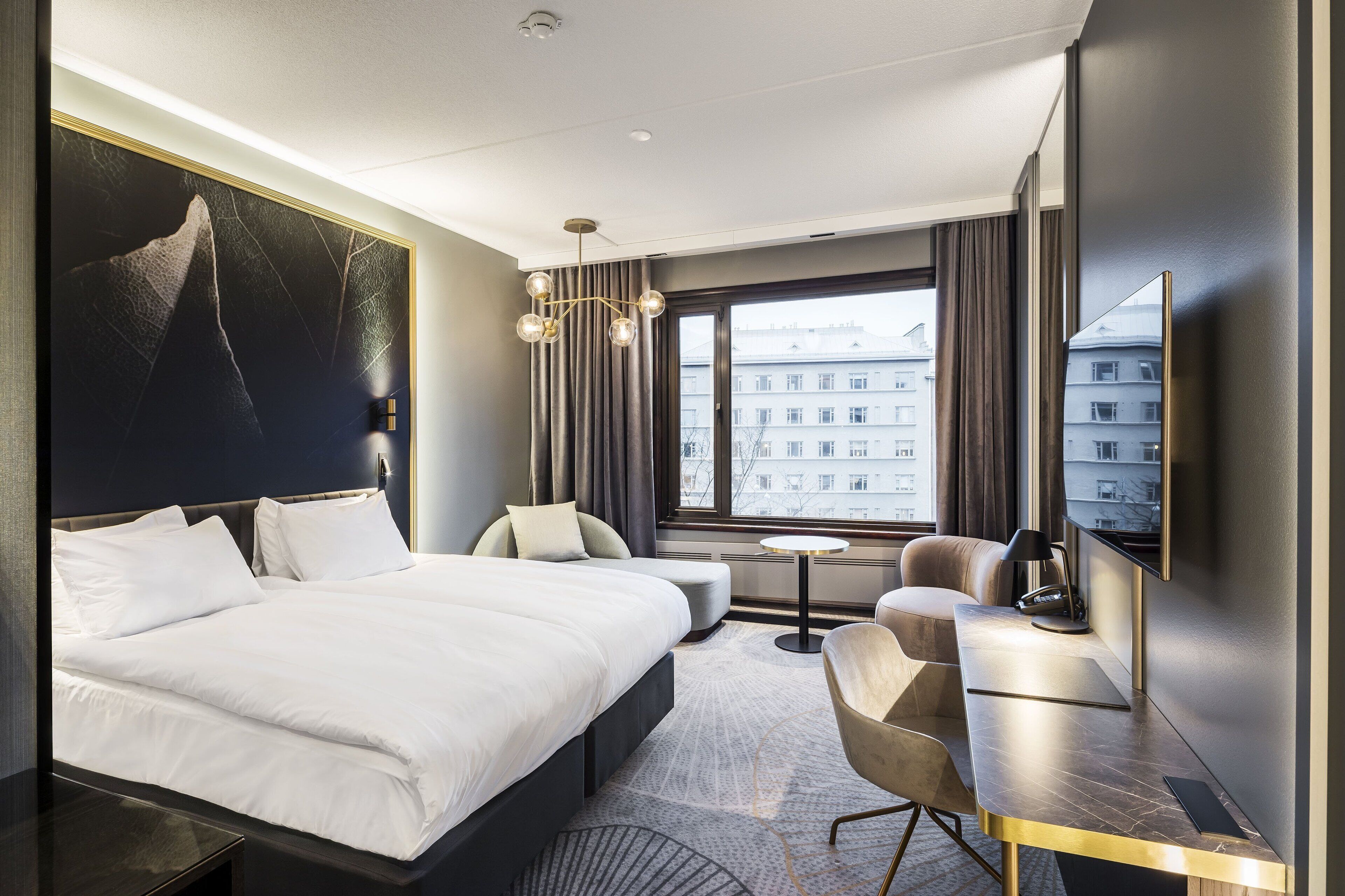 A modern hotel room with a large bed, a desk with a chair, a small round table, and a large window offering a city view.