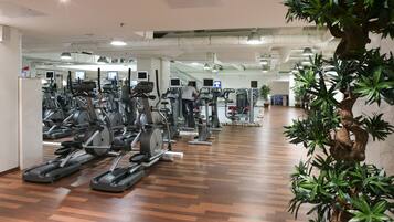 Una palestra con diverse tapis roulant e macchinari per esercizi, una persona che utilizza un macchinario, e una grande pianta da interno.