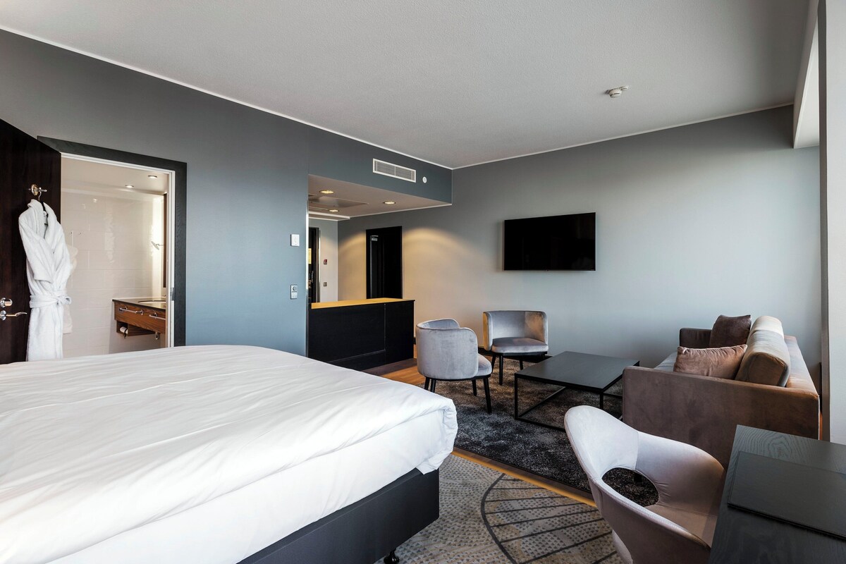 A modern hotel room with a large bed, a desk, a sofa, and a TV mounted on the wall.