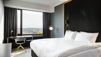 A modern hotel room with a large bed, a desk, and a chair. There is a window with curtains, a round table lamp, and a view of the city.