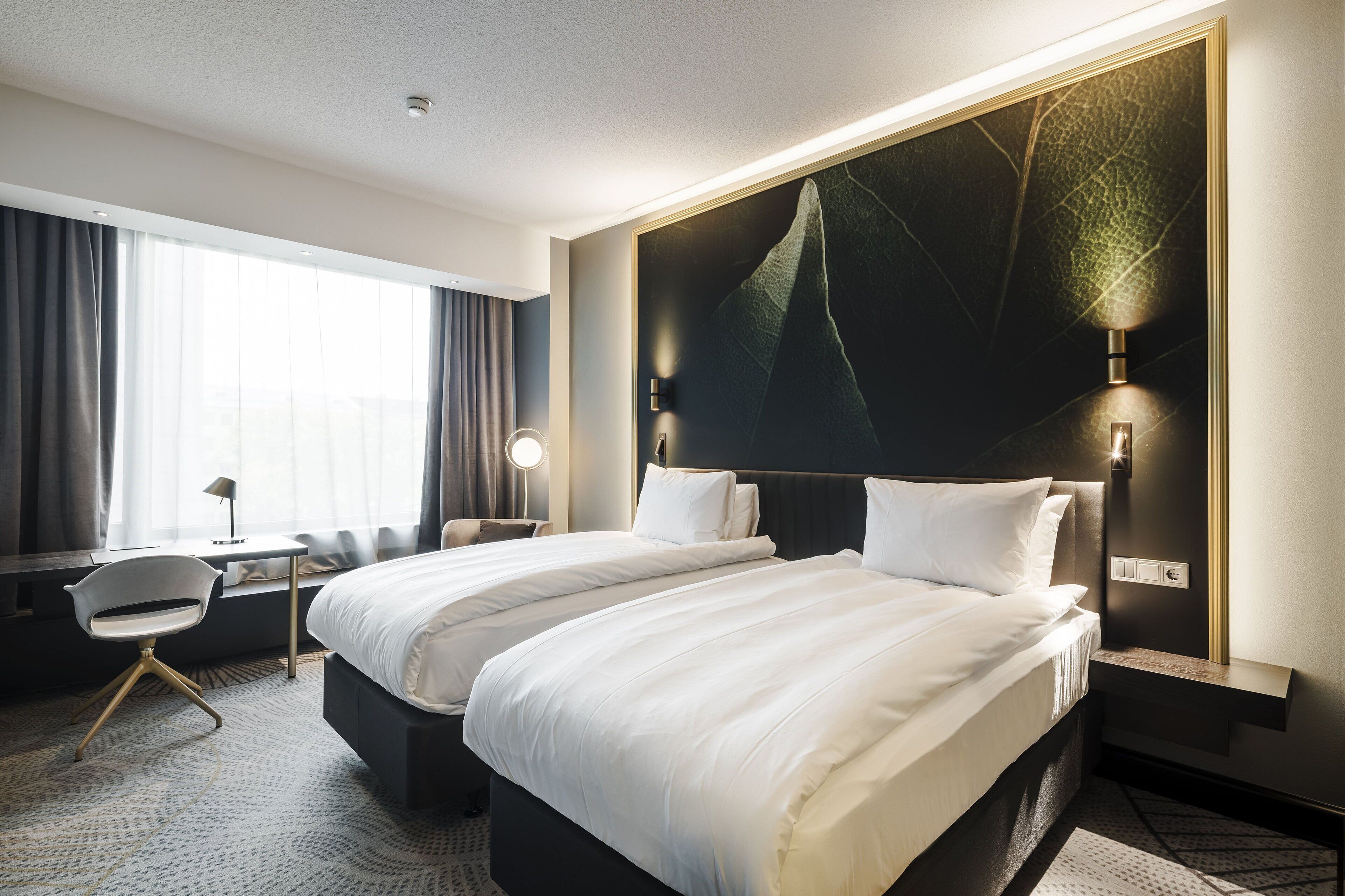 A hotel room with two beds, a desk, a chair, and a large window with curtains.