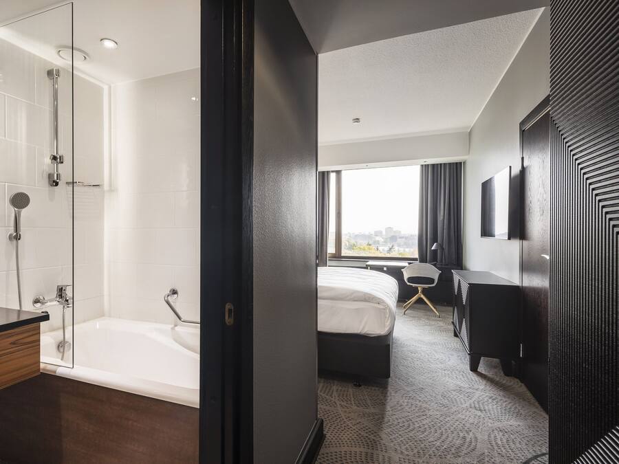 Quarto de hotel moderno com chuveiro em vidro, uma cama grande, mesa de cabeceira e vista da cidade.