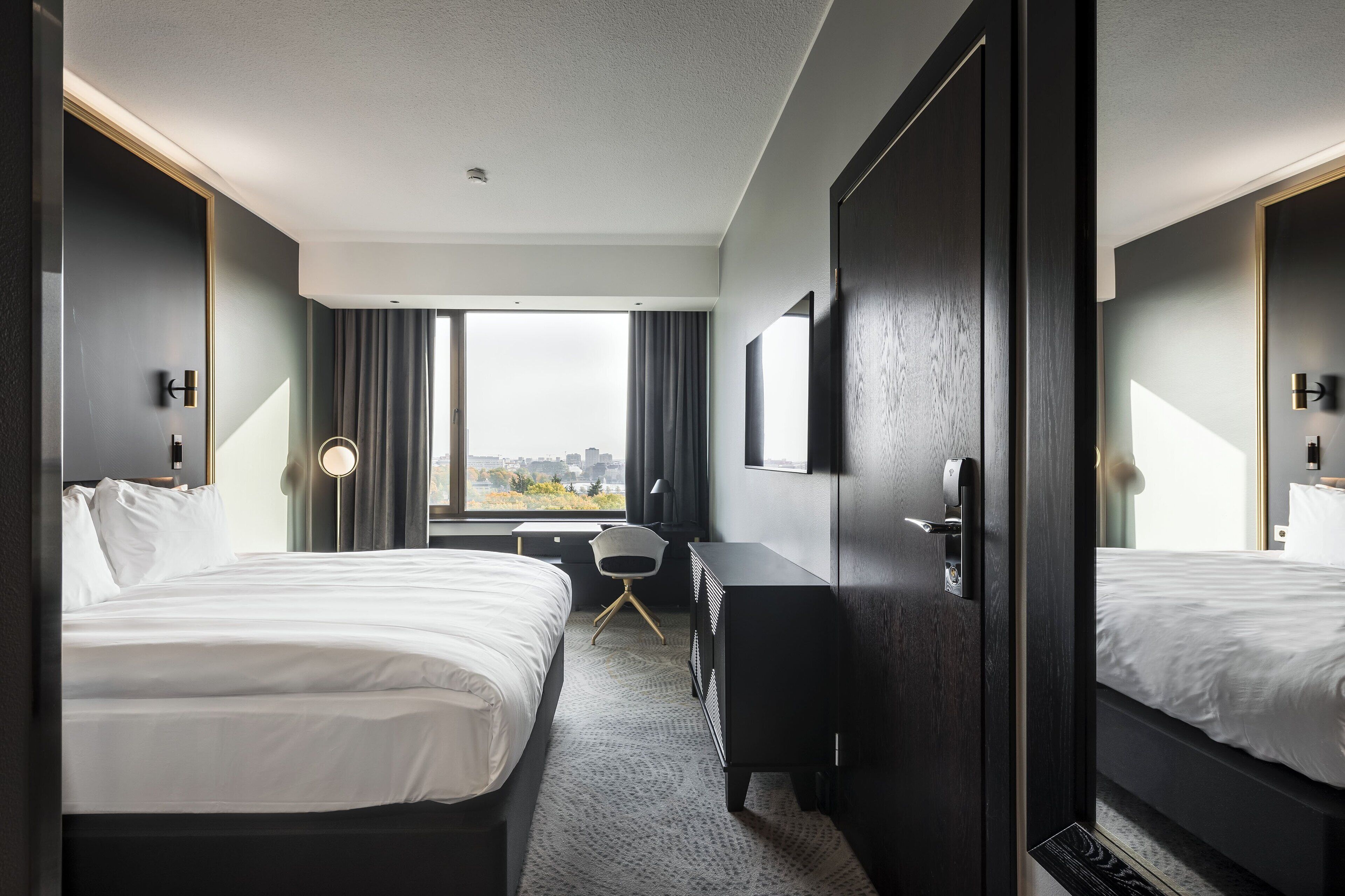 A hotel room with two beds, a desk, a chair, and a large window offering a city view.