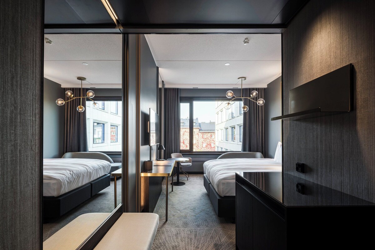 A modern hotel room with two beds, a desk, and a large window offering a city view.