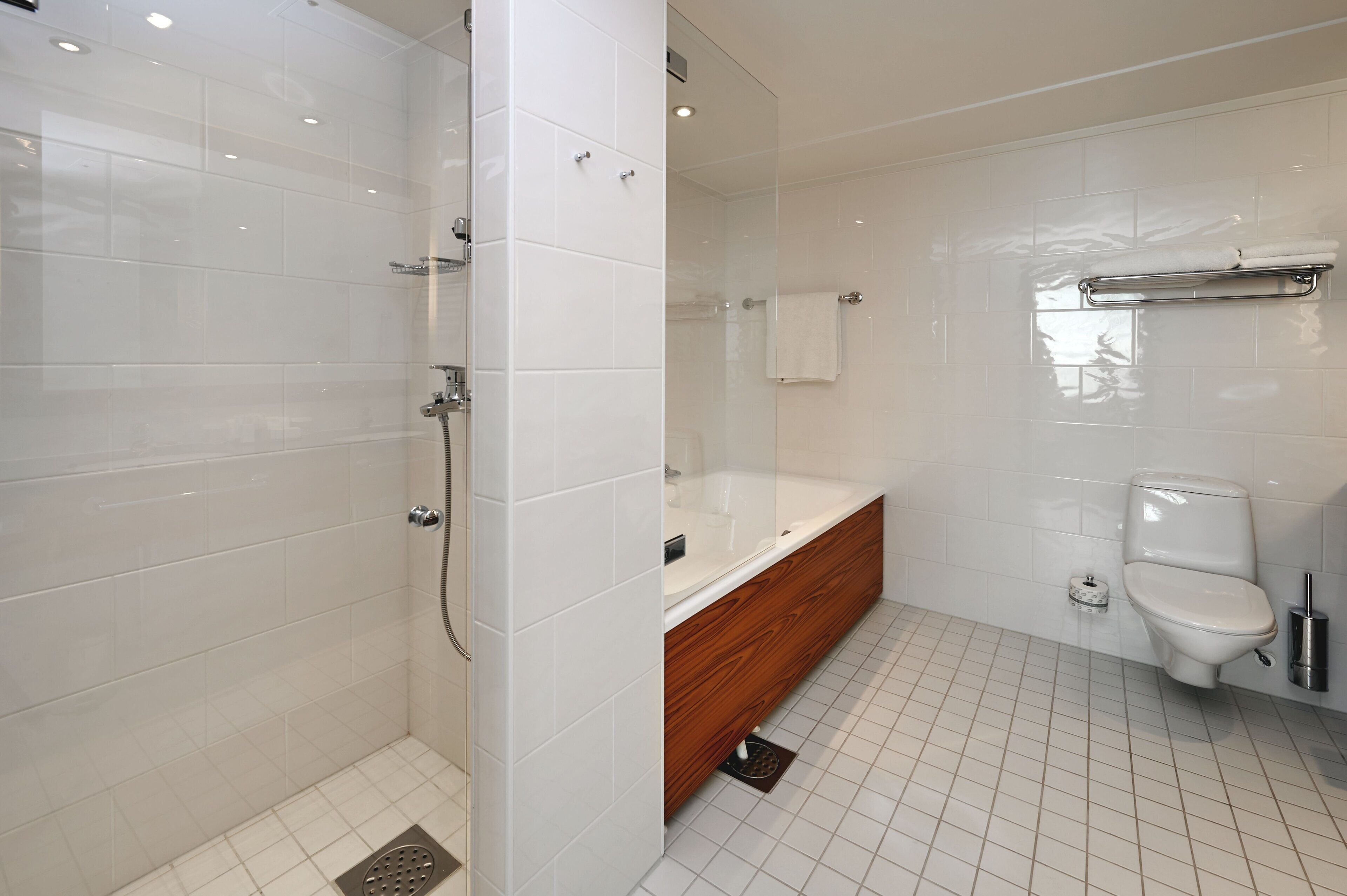 Une salle de bain moderne avec une douche à l’italienne, un meuble-lavabo en bois, des toilettes blanches et une baignoire entourée de parois en verre.