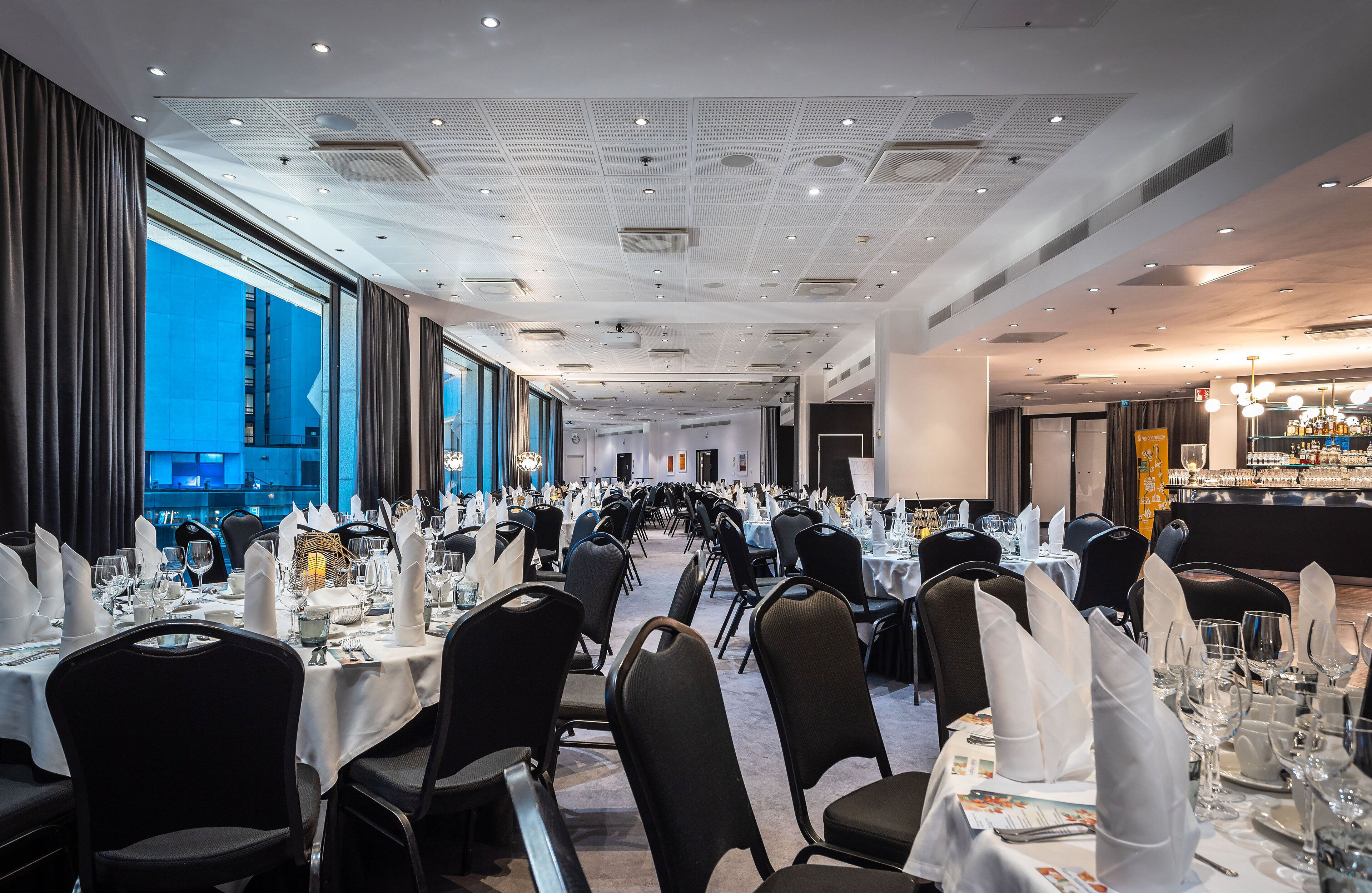 Een balzaal met rijen tafels gedekt voor een formeel evenement, grote ramen aan één kant en een bar aan de andere kant.