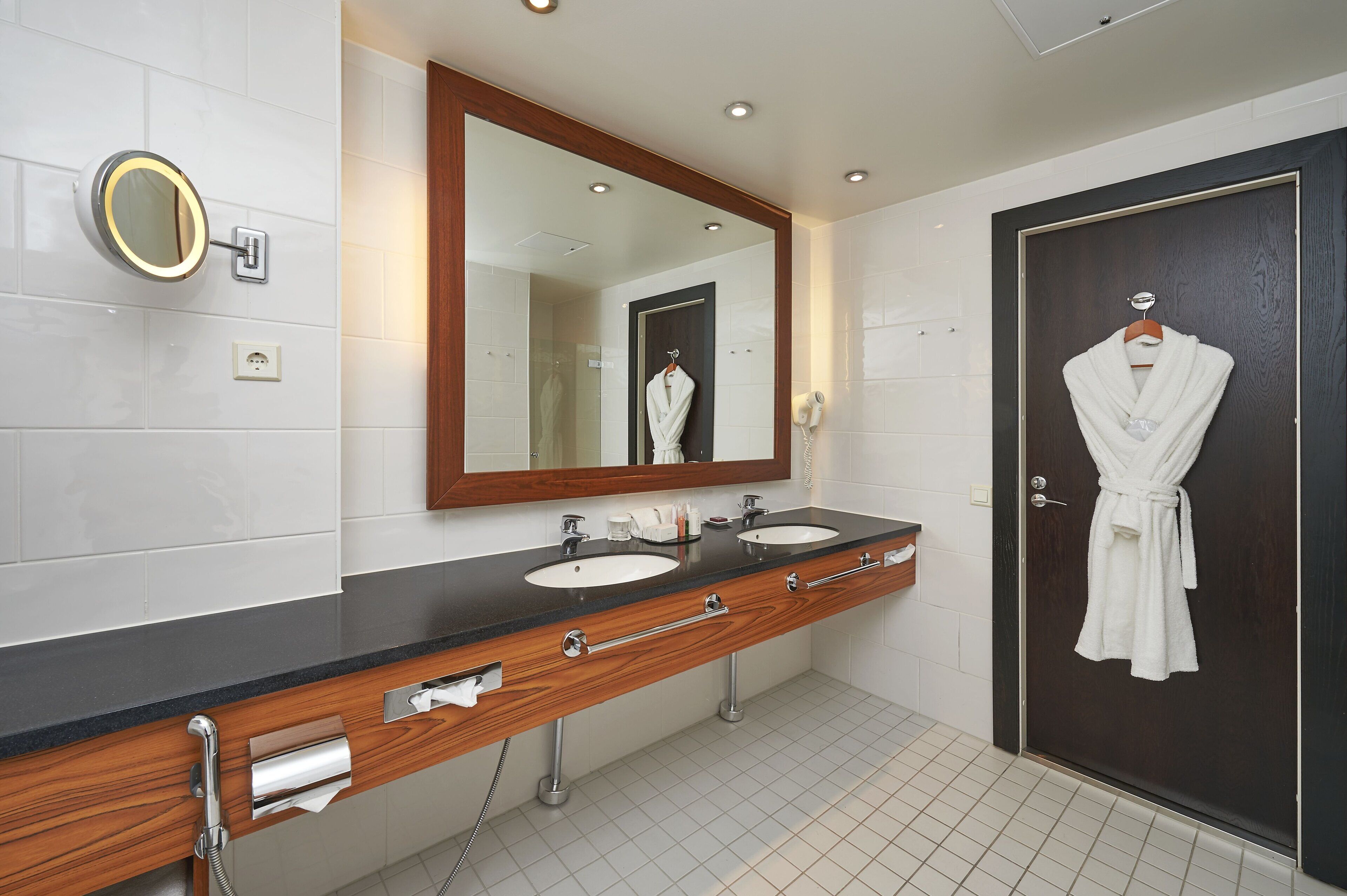 A bathroom with two sinks, a large mirror, a robe hanging on the door, and a towel rack.