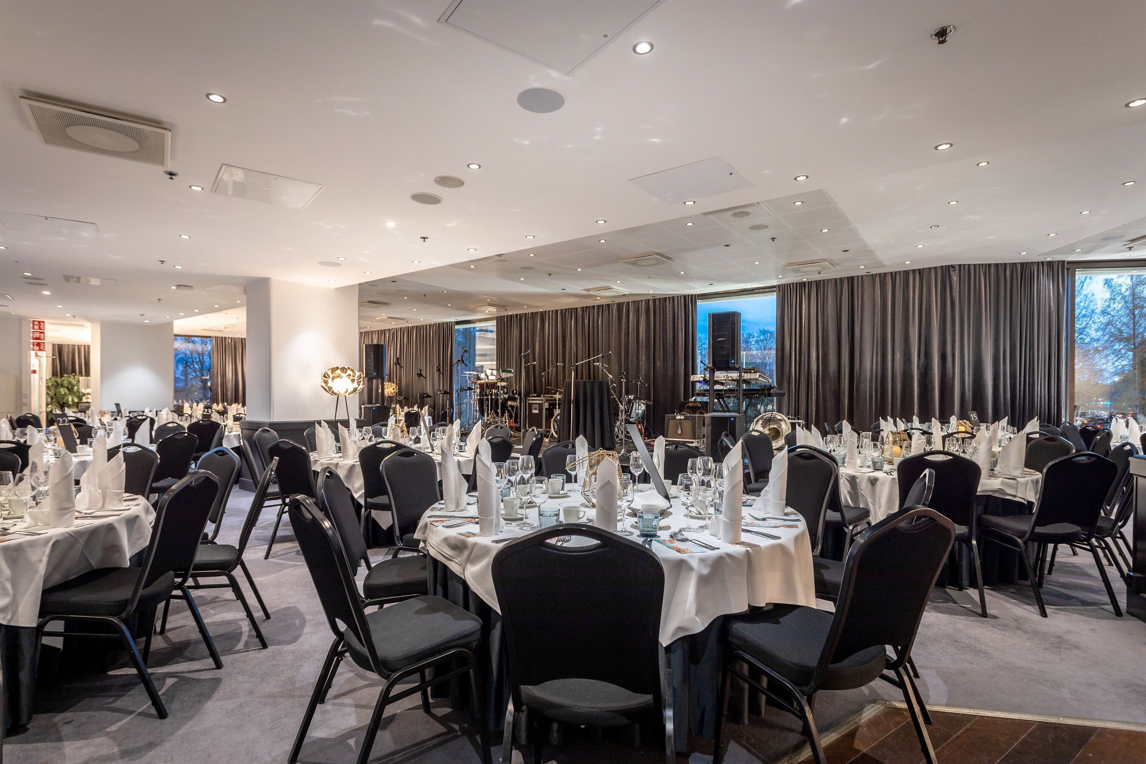 Un salón de banquetes con mesas redondas dispuestas para comer, un escenario con instrumentos musicales y un amplio ventanal con cortinas.