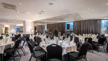 Un salón de banquetes con mesas redondas dispuestas para comer, un escenario con instrumentos musicales y un amplio ventanal con cortinas.