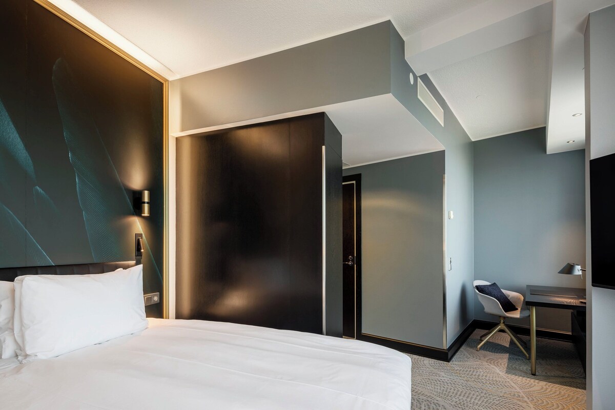 A modern hotel room with a large bed, a desk, and a chair. There is a wardrobe and a television.