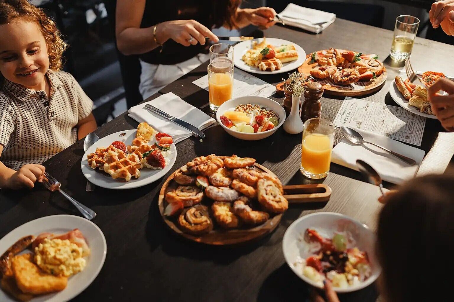 A group of people dining at a table with various dishes, including waffles and a fruit salad.