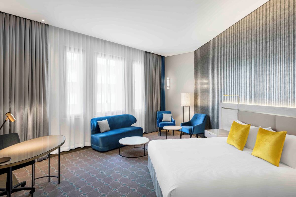 A hotel room with a large bed, two blue armchairs, a small round table, a desk with a lamp, and a large window with curtains.