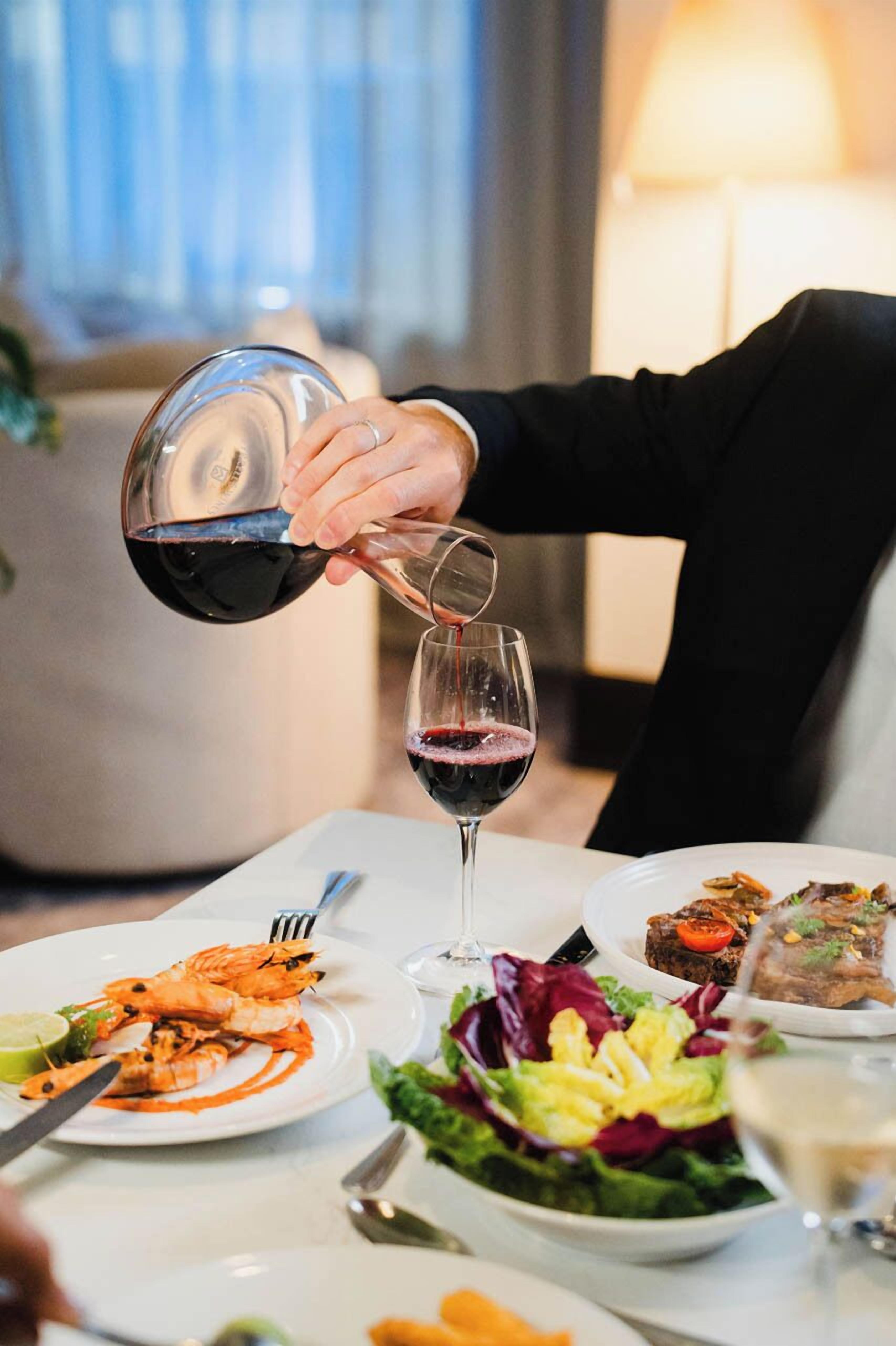 A person pouring red wine into a glass, a plate of seafood, and a salad on a table.