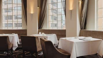 Ein Hotelrestaurant mit gedeckten Tischen, großen Fenstern mit Vorhängen und Blick auf die Stadt.
