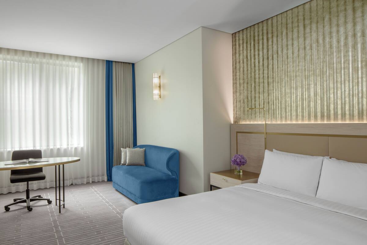 A hotel room with a bed, a desk, a blue armchair, a nightstand, and a window with curtains.
