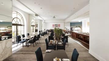 A spacious dining area with high ceilings, large windows, and a central plant. Tables are neatly arranged with chairs, and a buffet counter is visible on the right.