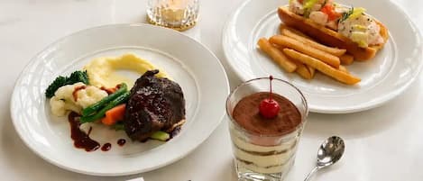 A dining table with plates of food, a dessert, and a glass of dessert with a cherry on top.