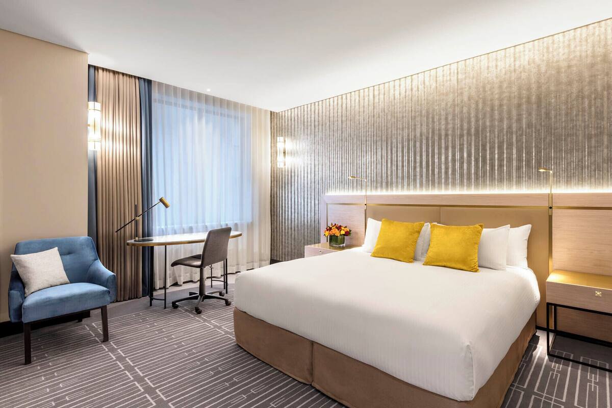 A hotel room with a large bed, a desk, a chair, and a nightstand with a lamp.
