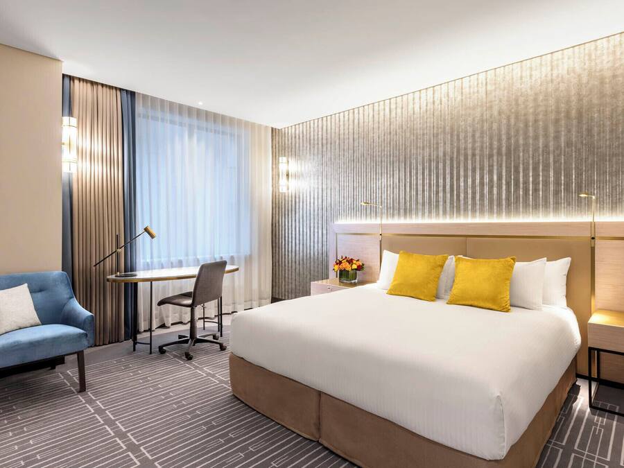 Quarto de hotel com uma cama grande, uma escrivaninha, uma cadeira e uma mesa de cabeceira com abajur.