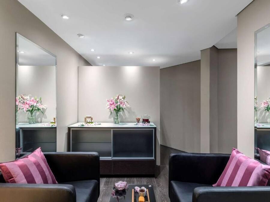 A modern spa room with a large mirror, two black sofas with pink pillows, a reception desk with a vase of flowers, and a wooden tray with a candle and a small vase.