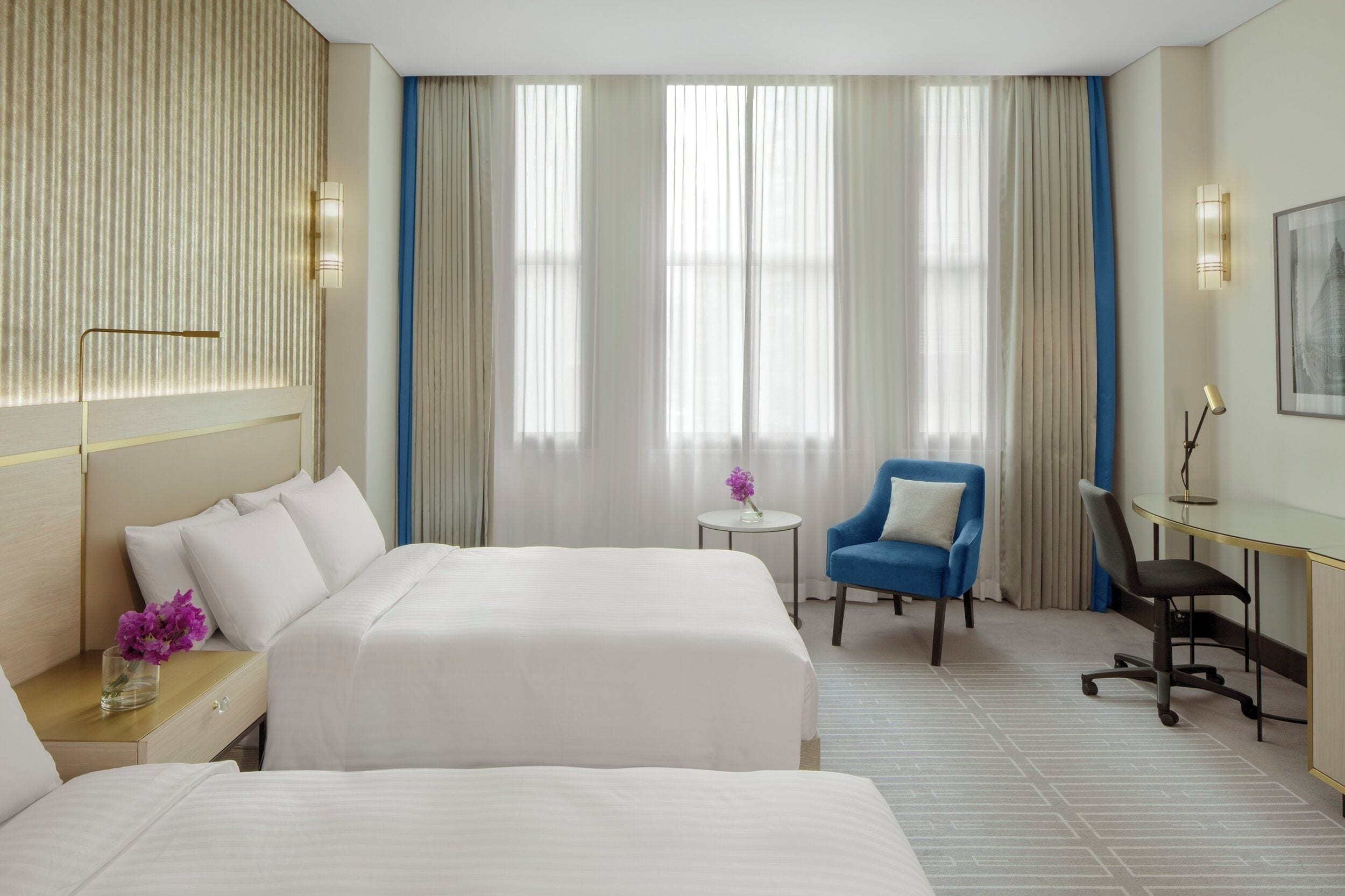 A hotel room with two beds, a desk, a chair, and a small table with a vase of flowers.
