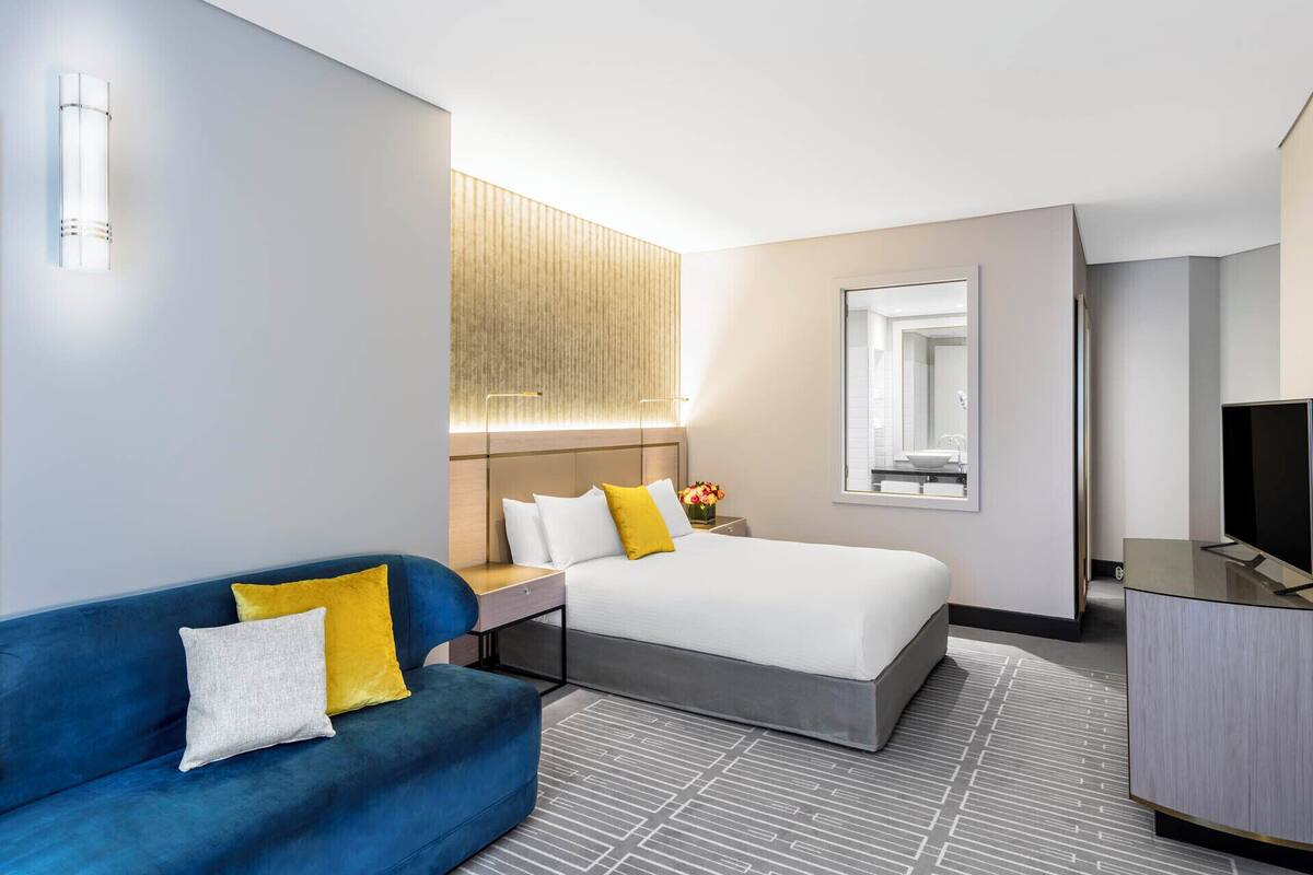 A hotel room with a bed, a blue sofa, a desk with a lamp, a TV, and a large window.