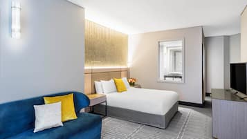 A hotel room with a bed, a blue sofa, a desk with a lamp, a TV, and a large window.