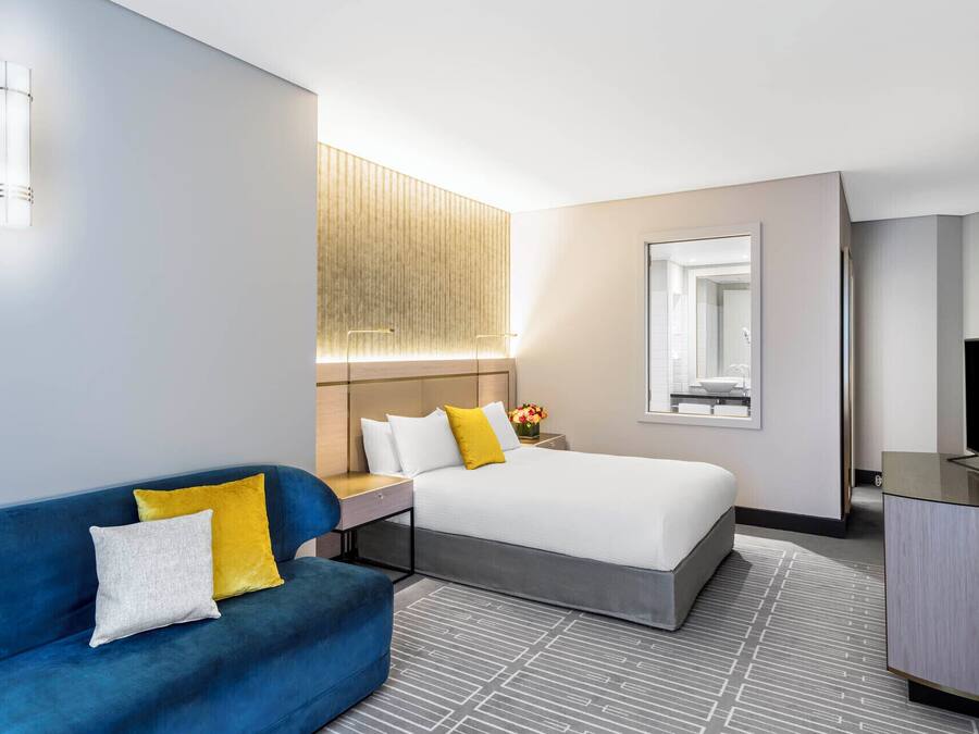 A hotel room with a bed, a blue sofa, a desk with a lamp, a TV, and a large window.