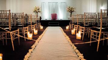 Preparação para uma cerimônia de casamento com tapete branco no corredor, cadeiras de madeira, velas e arranjos florais.