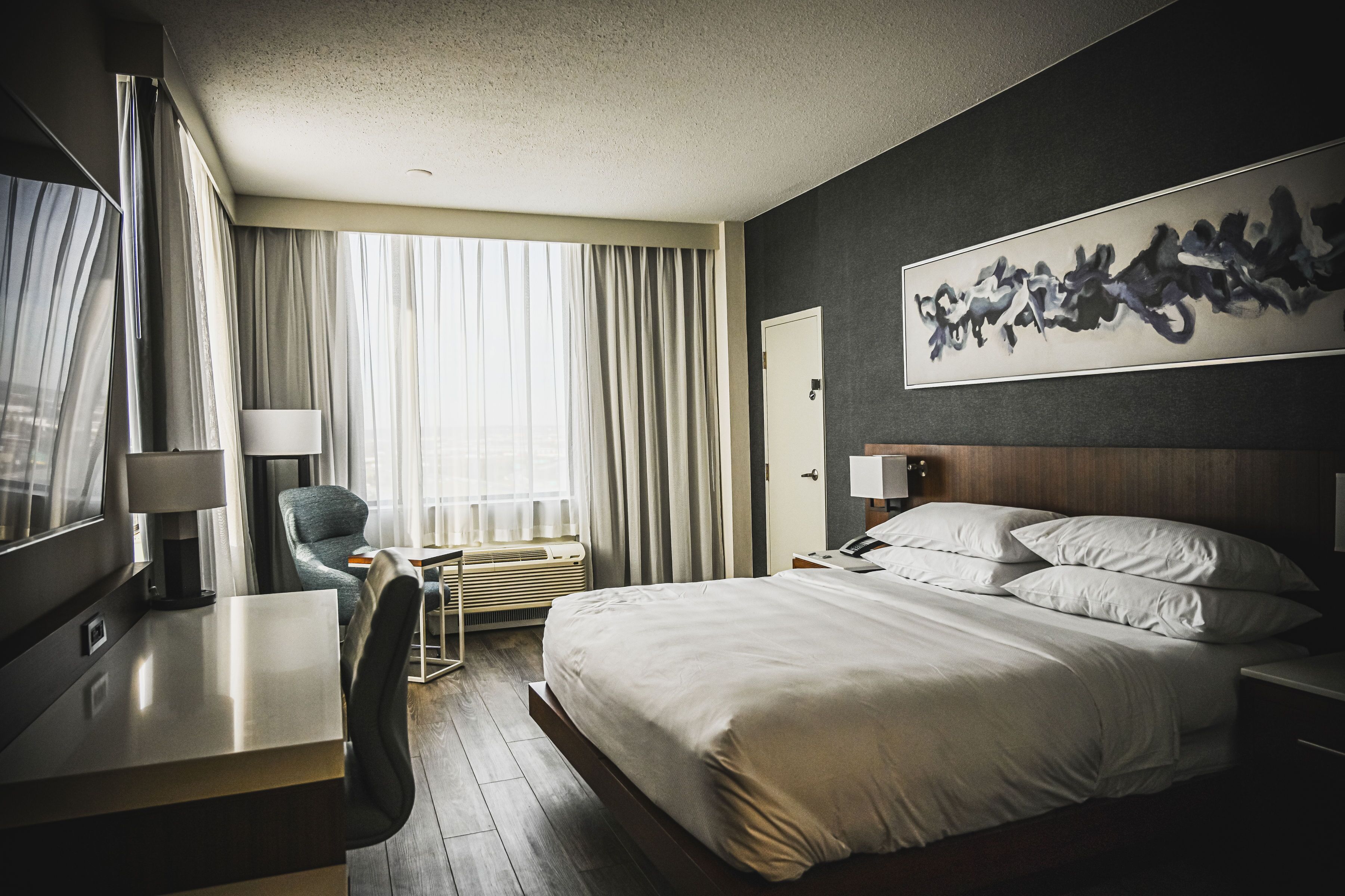 A hotel room with a bed, a desk, a chair, a large window with curtains, a wall art piece, and a door.