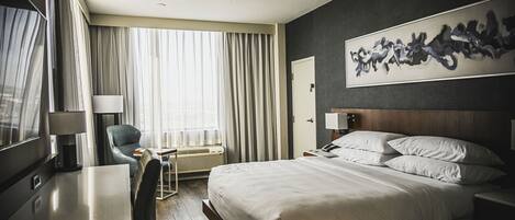 A hotel room with a bed, a desk, a chair, a large window with curtains, a wall art piece, and a door.