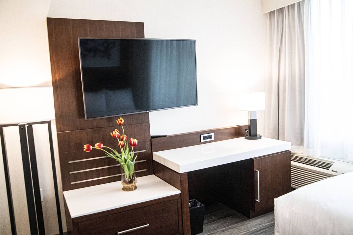 Une chambre d’hôtel dotée d’un téléviseur à écran plat fixé au mur, d’une table de chevet blanche ornée d’un vase de tulipes, d’un bureau en bois avec une poubelle et d’un lit recouvert de draps blancs.