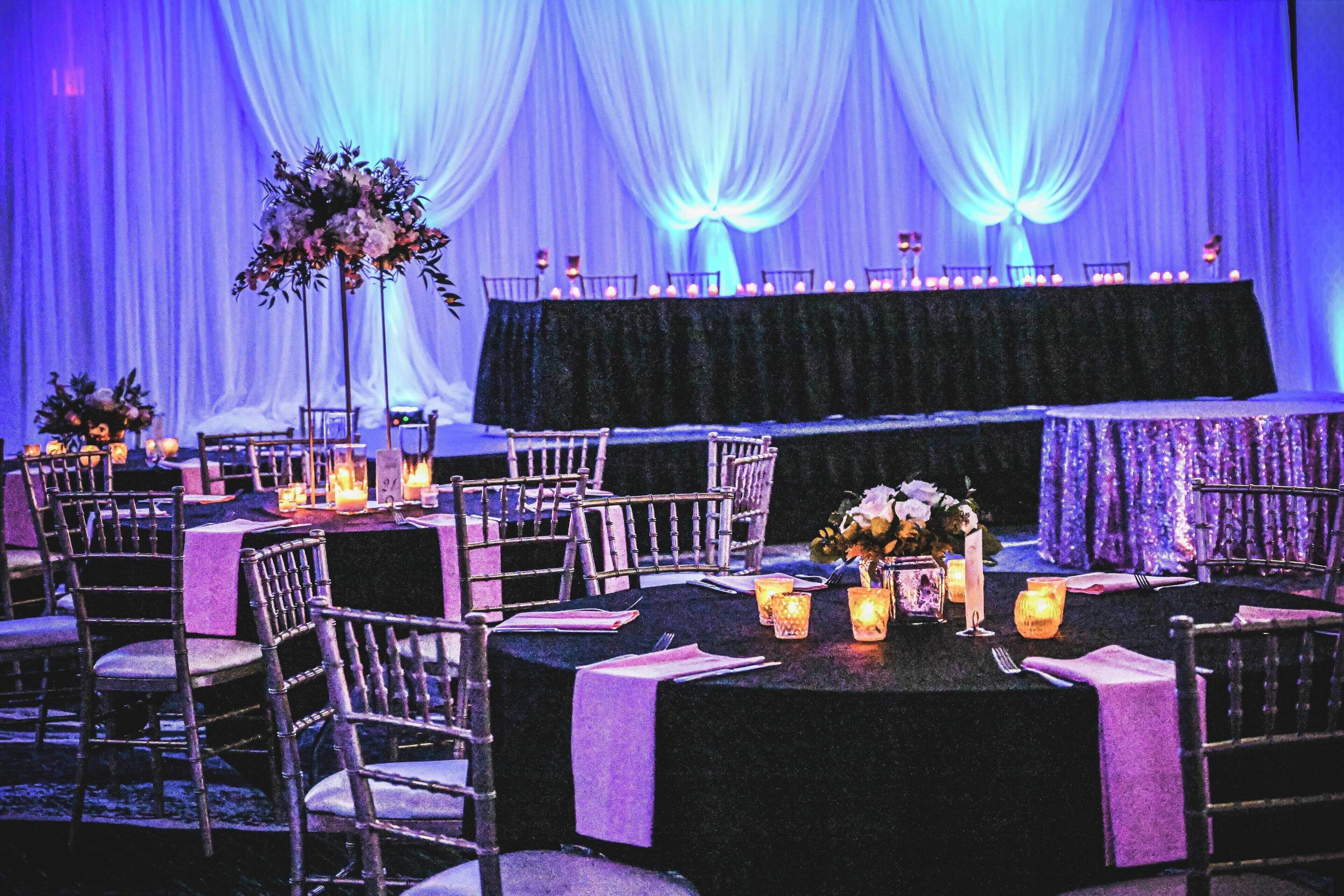 Un salón de banquetes con mesas redondas, sillas y un escenario con cortinas.