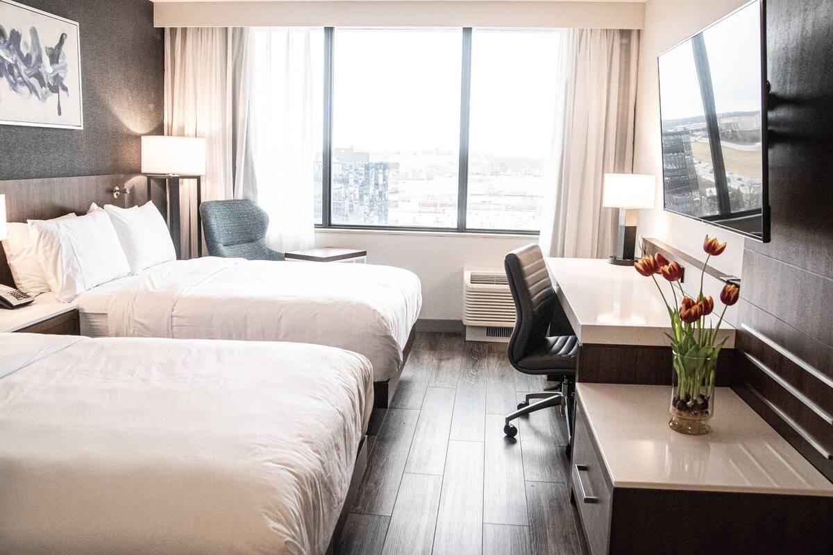 Une chambre d’hôtel avec deux lits, un bureau, une chaise, un vase avec des fleurs, une télévision et une fenêtre donnant sur la ville.
