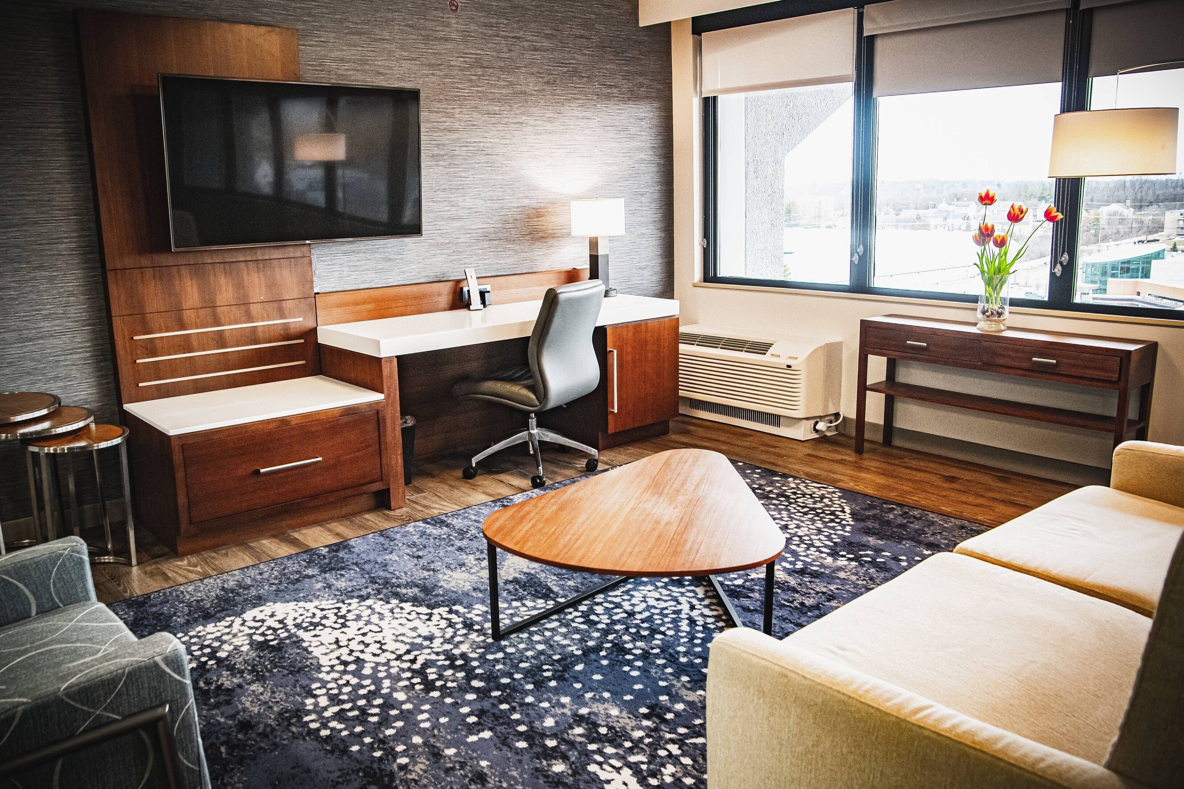 A modern hotel room with a wooden desk, a flat-screen TV, a comfortable sofa, and a large window with a view.