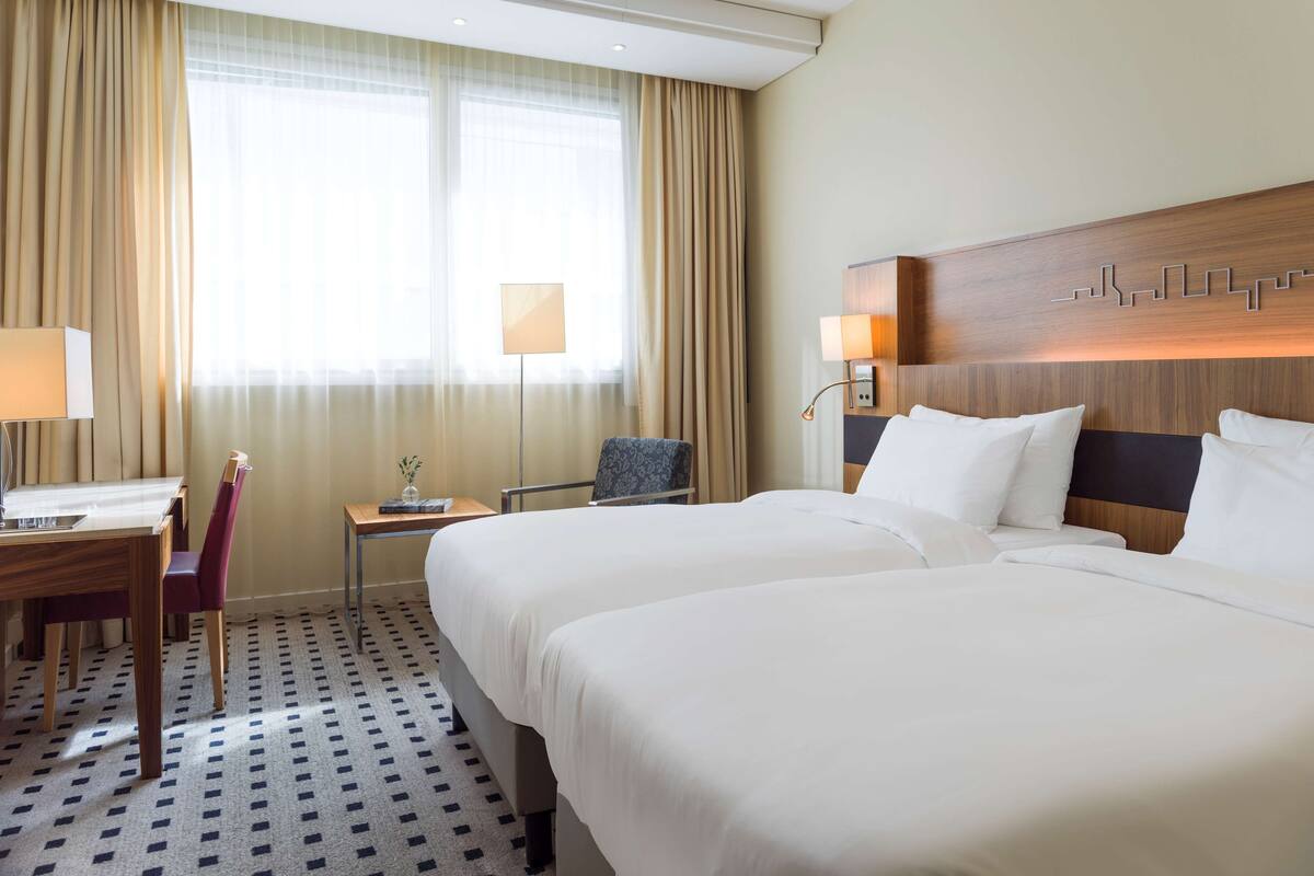 A hotel room with two beds, a desk, a chair, and a small table with a lamp.