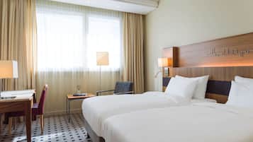 A hotel room with two beds, a desk, a chair, and a small table with a lamp.