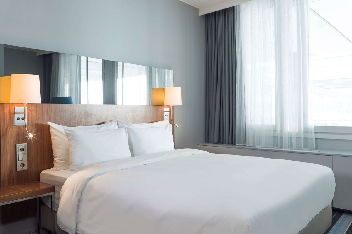 Habitación de hotel con una cama grande, dos lámparas de noche, una mesita de noche, una ventana con cortinas y un espejo sobre la cama.