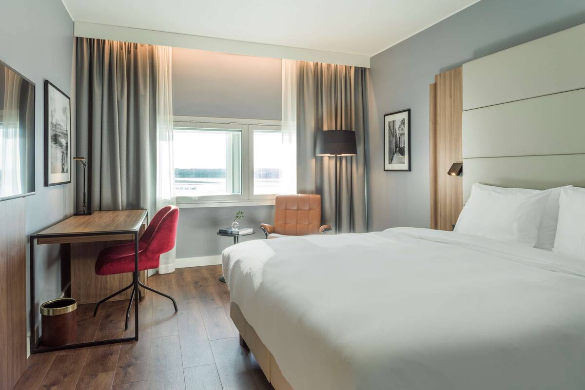 A hotel room with a large bed, a desk, a red chair, a small table with a vase, and a window with curtains.