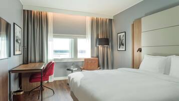 Quarto de hotel com uma cama grande, uma escrivaninha, uma cadeira vermelha, uma mesinha com um vaso e uma janela com cortinas.