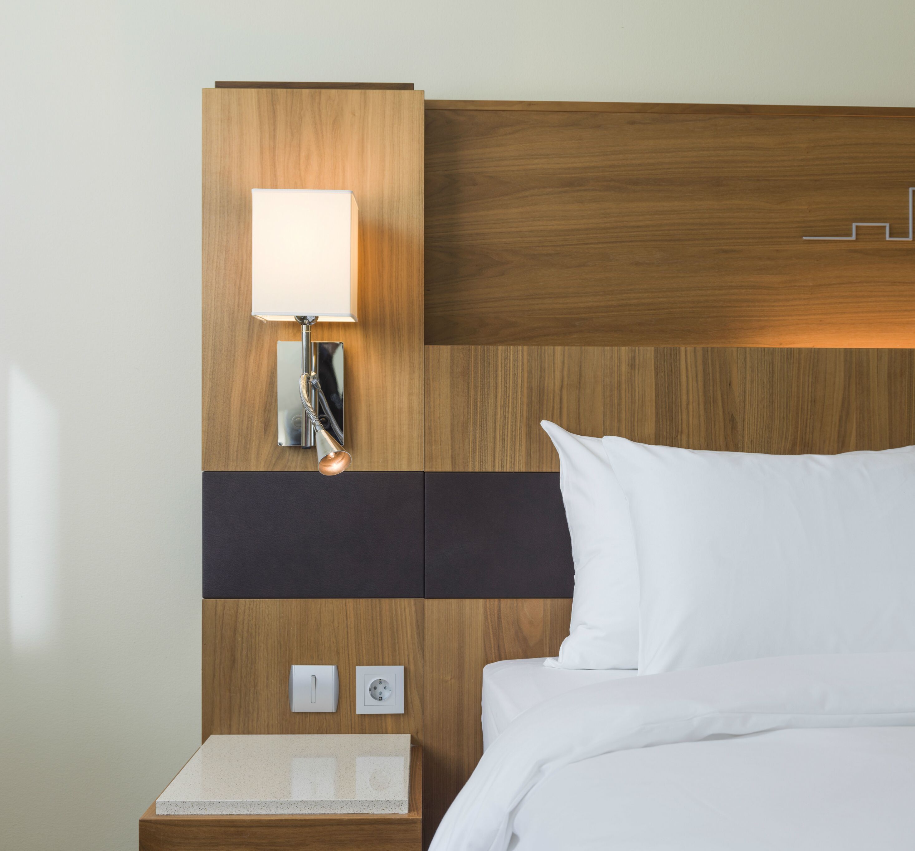 A hotel room with a wooden headboard, a bedside table with a lamp, a power outlet, and a small shelf.