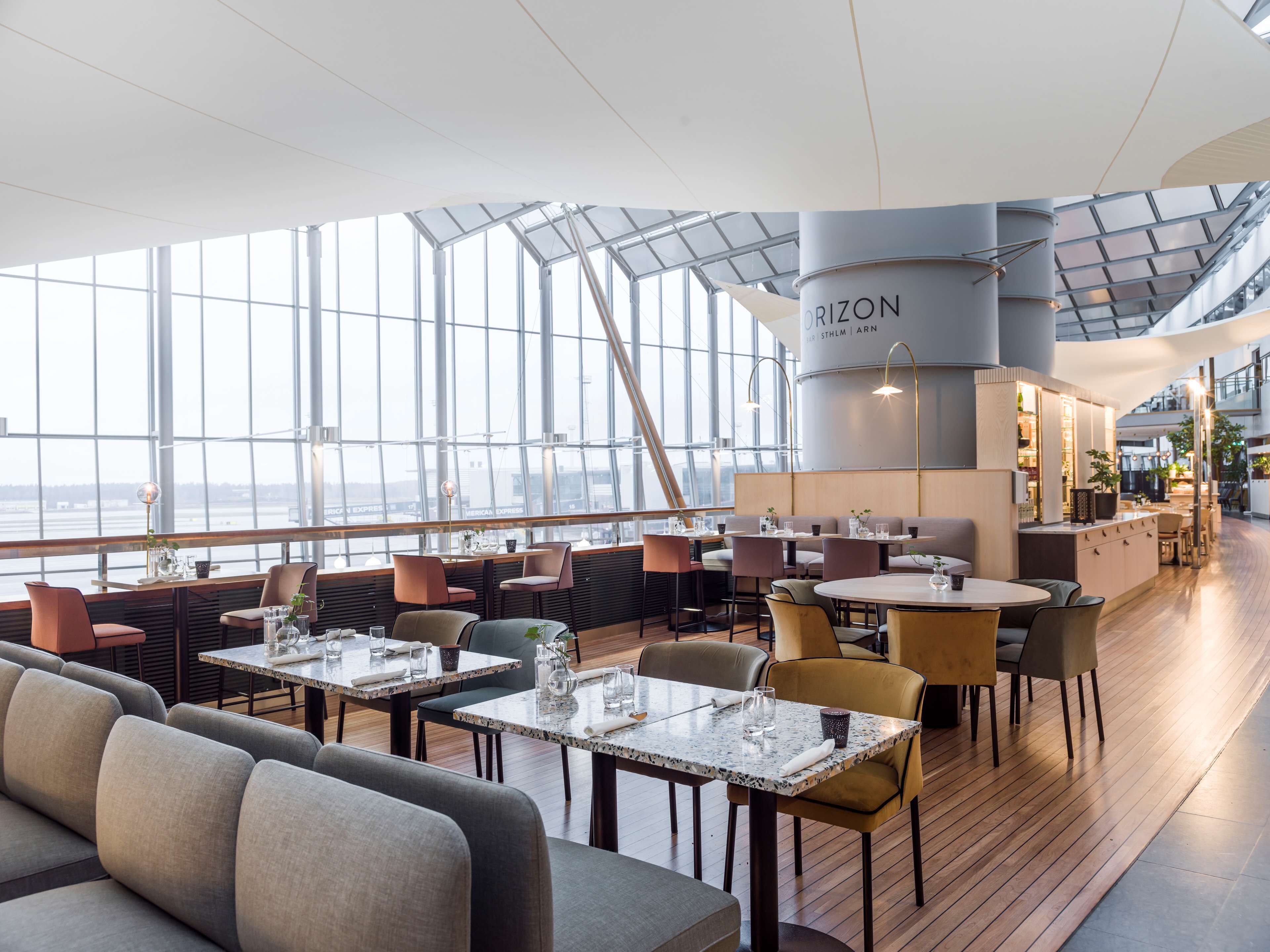 A modern restaurant with a high ceiling, large windows, and a view of the water. Tables and chairs are arranged for dining.