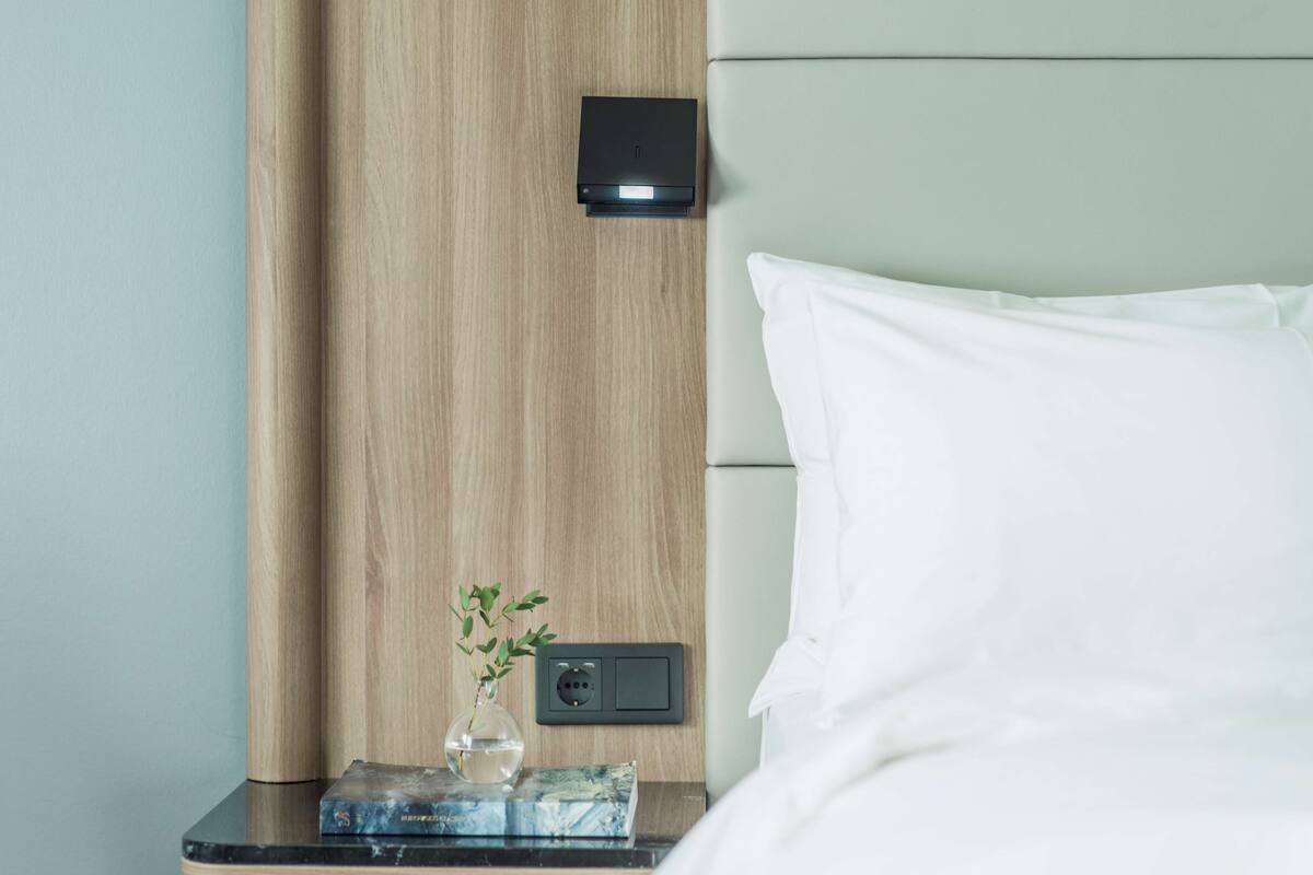 A neatly made bed with white sheets, a wooden headboard, a bedside table with a vase of green plants, and a small black device on the wall.